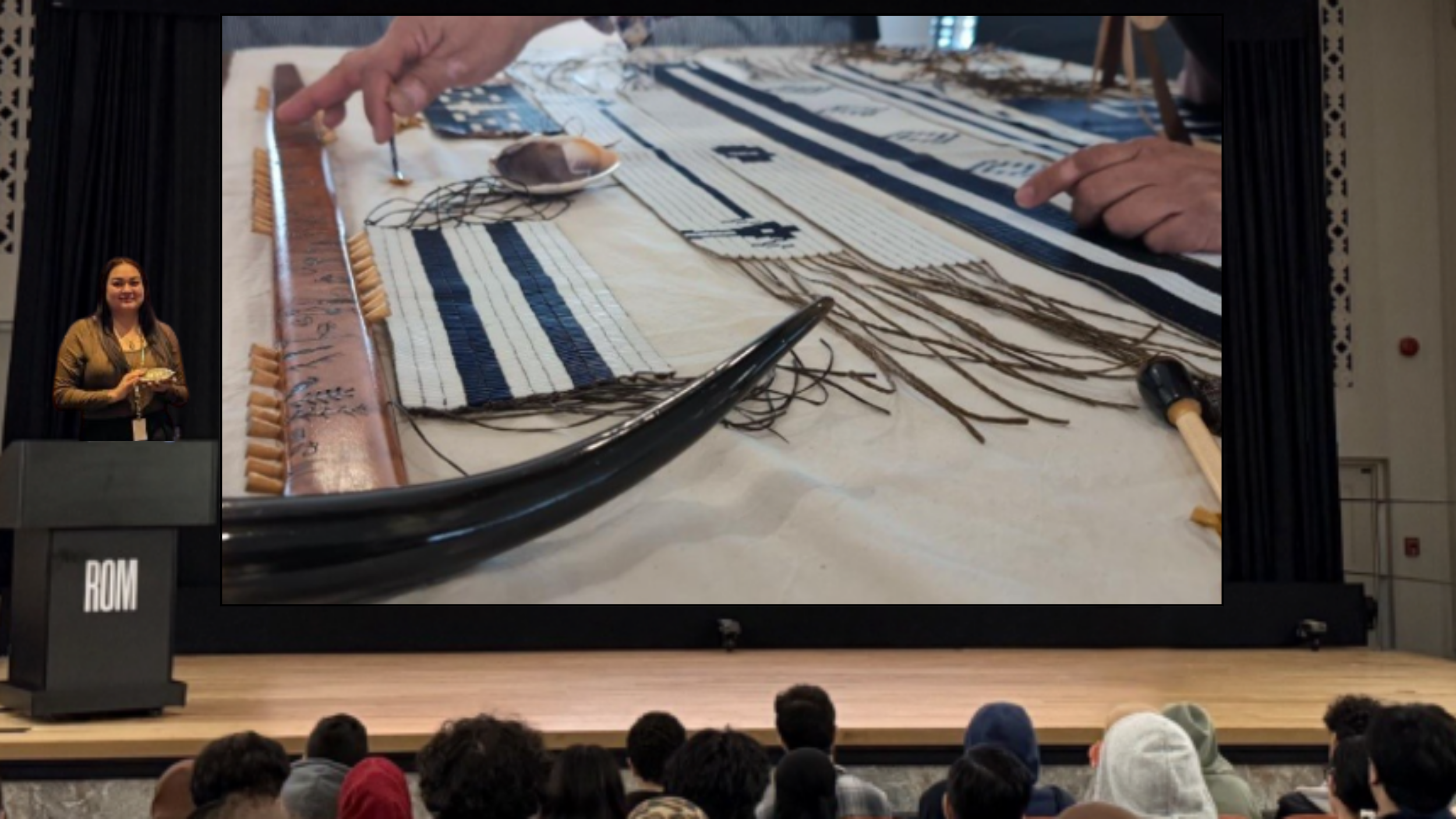 An Indigenous woman stands at a podium on a stage in front of a group of young people, gesturing to wampum belts and other belongings displayed on a screen.