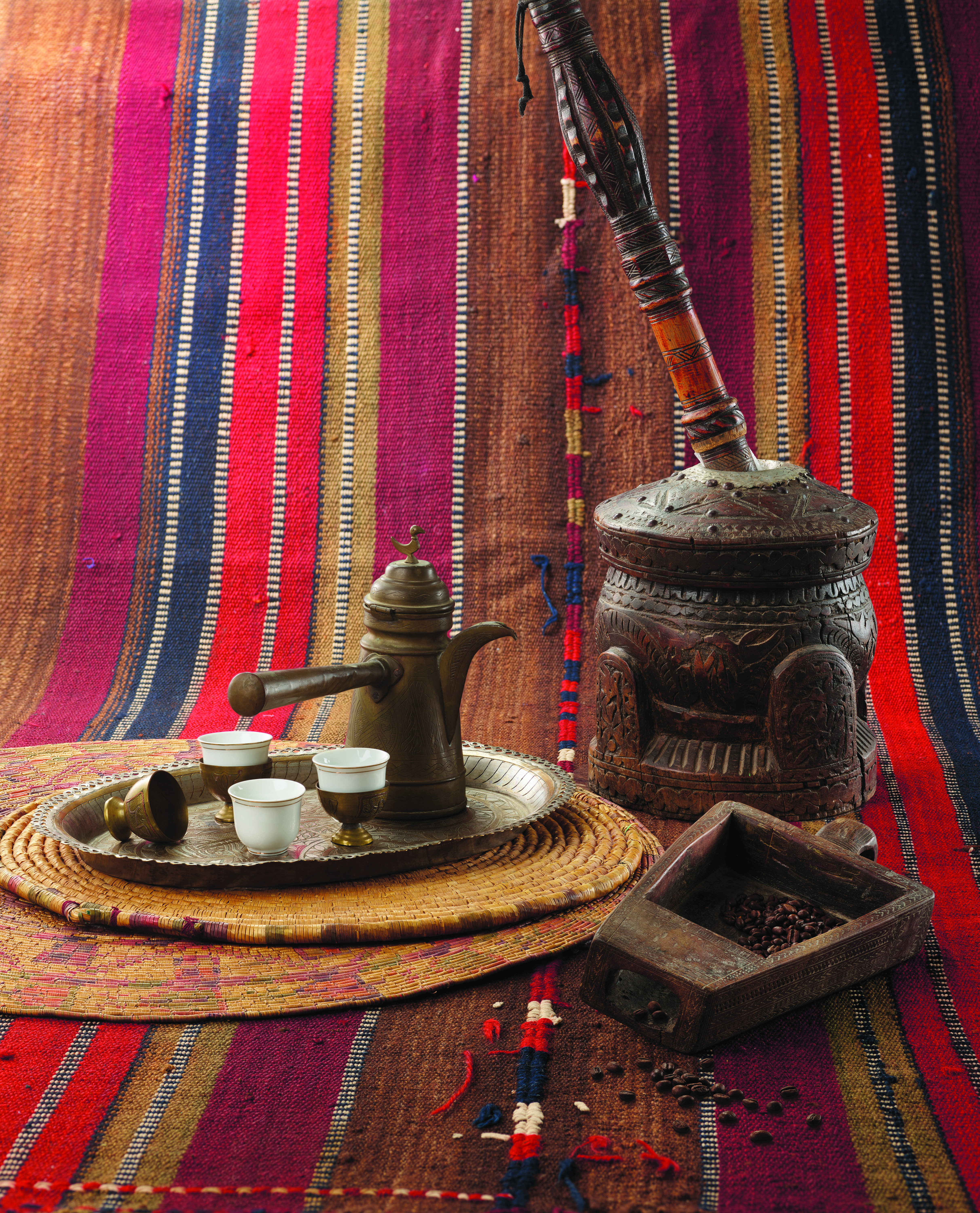 Traditional coffee-serving set with a metal pot and small cups on a tray, alongside carved wooden containers and coffee beans on a colourful striped textile backdrop.