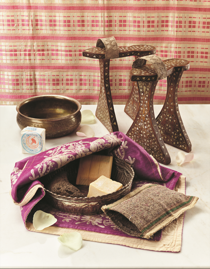 Traditional grooming items arranged on embroidered fabric, including carved wooden platform sandals, a metal basin, soap, a comb, and woven textiles.