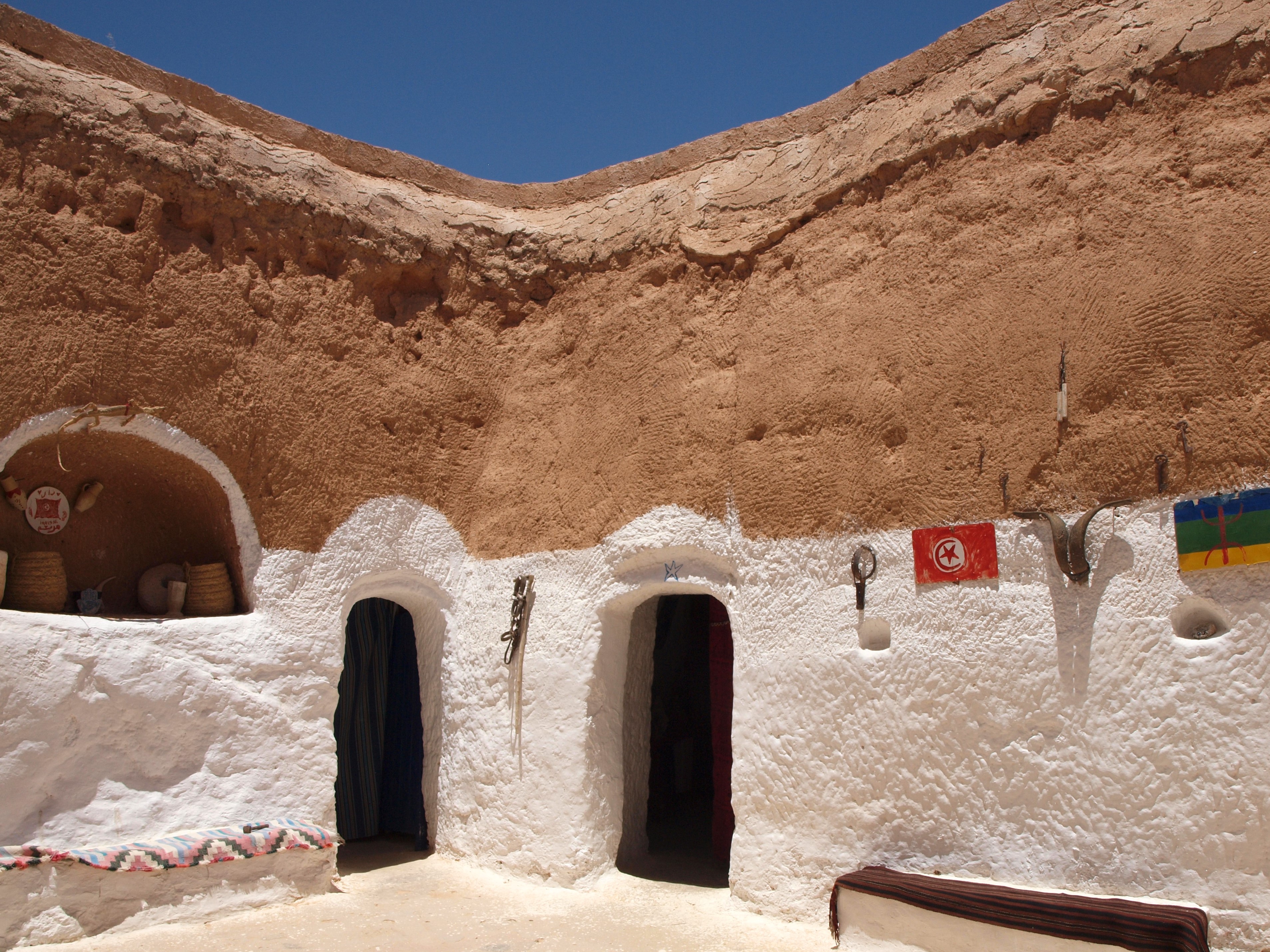 A picturesque, weathered white cave dwelling under a clear blue sky, with colourful flags and decorations on the wall.