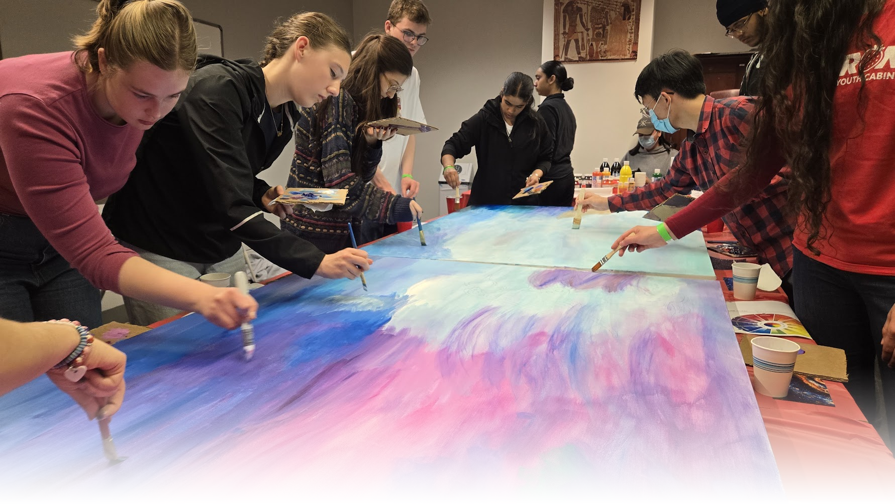 A group of Youth Cabinet members work together to paint a canvas with shades of blue, purple and pink.