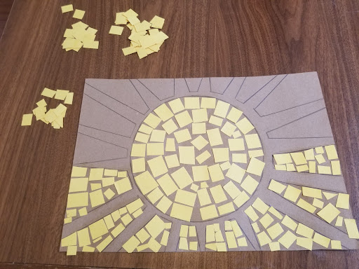 A sun design on cardboard sits on a table. The centre of the sun and half of the space between sun rays are filled with yellow squares. Additional piles of yellow squares sit on the table next to the design.
