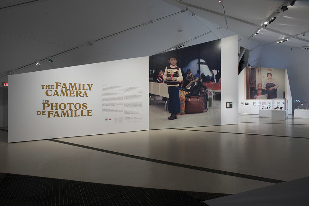The entrance to The Family Camera exhibition. A large wall displays the title “The Family Camera” in English and French, next to an enlarged image of a young boy standing in front of a pile of bags in the waiting area of an airport.