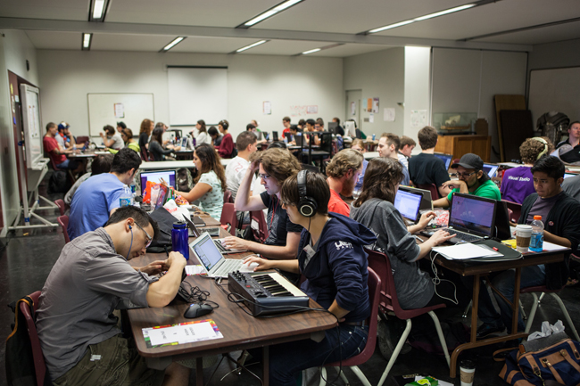 Game Jam participants sit hard at work in front of tables strewn with computers, caffeinated beverages and sketch books