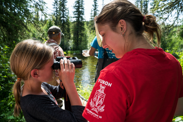 A student from the Yukon Youth Conservatio Corps helps a young bioblitz participant get a closer look downstream with binoculars. Photo by Stacey Lee Kerr