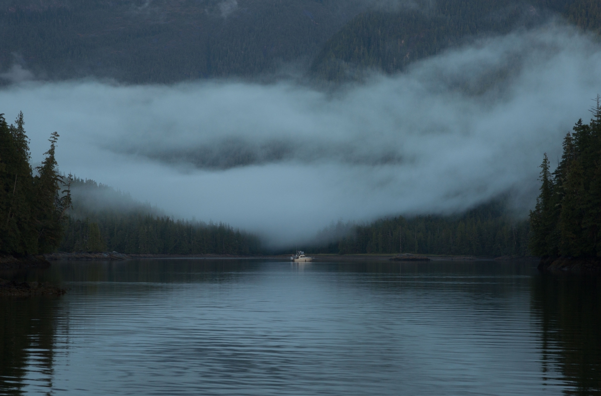 A boat sits on an inlet, with misty, tree covered hills rising in the background