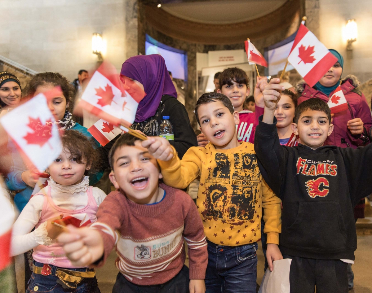 Un groupe d’enfants syriens souriant et agitant des drapeaux canadiens devant la Galerie Schad de la biodiversité du ROM.