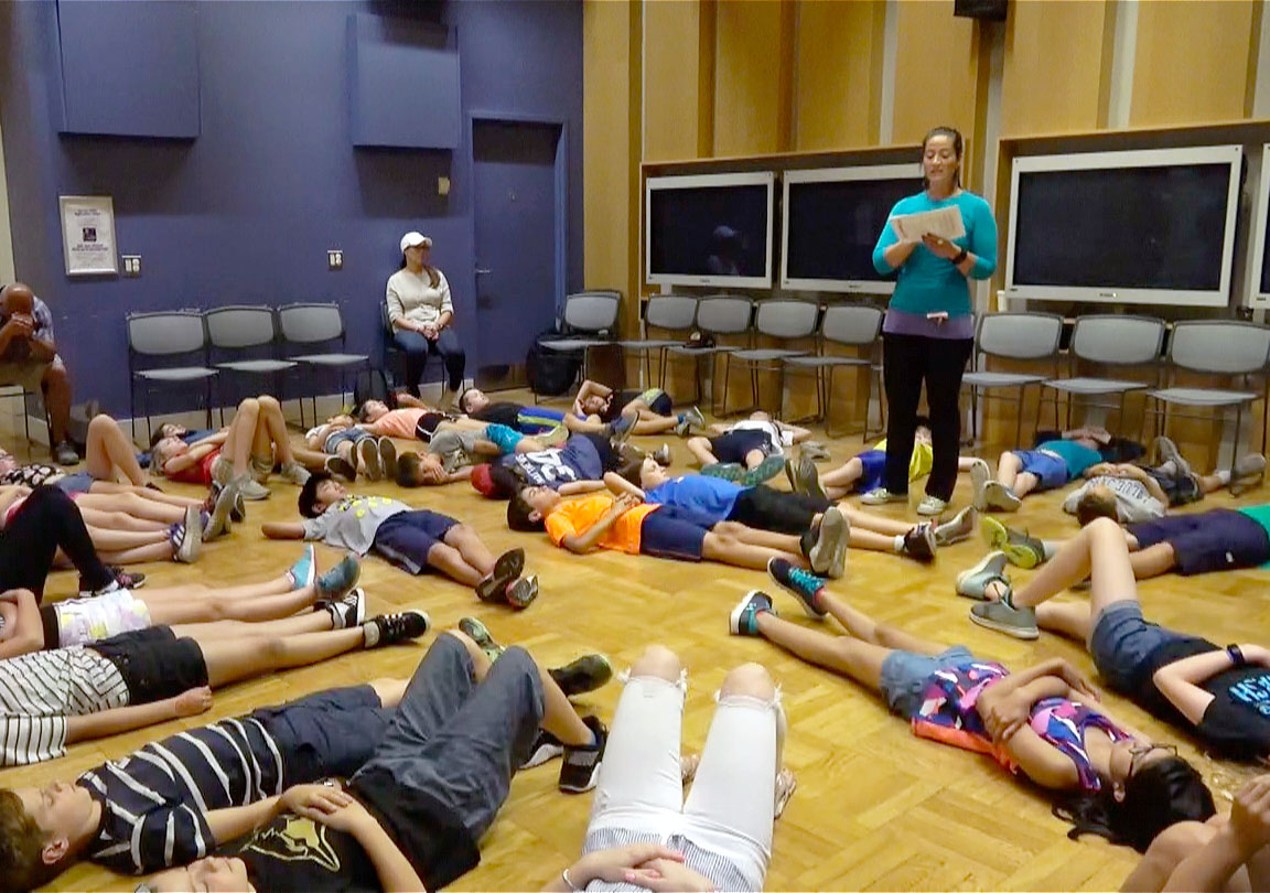 Students lie on the floor in a circle. A woman stands next to them, telling a story.