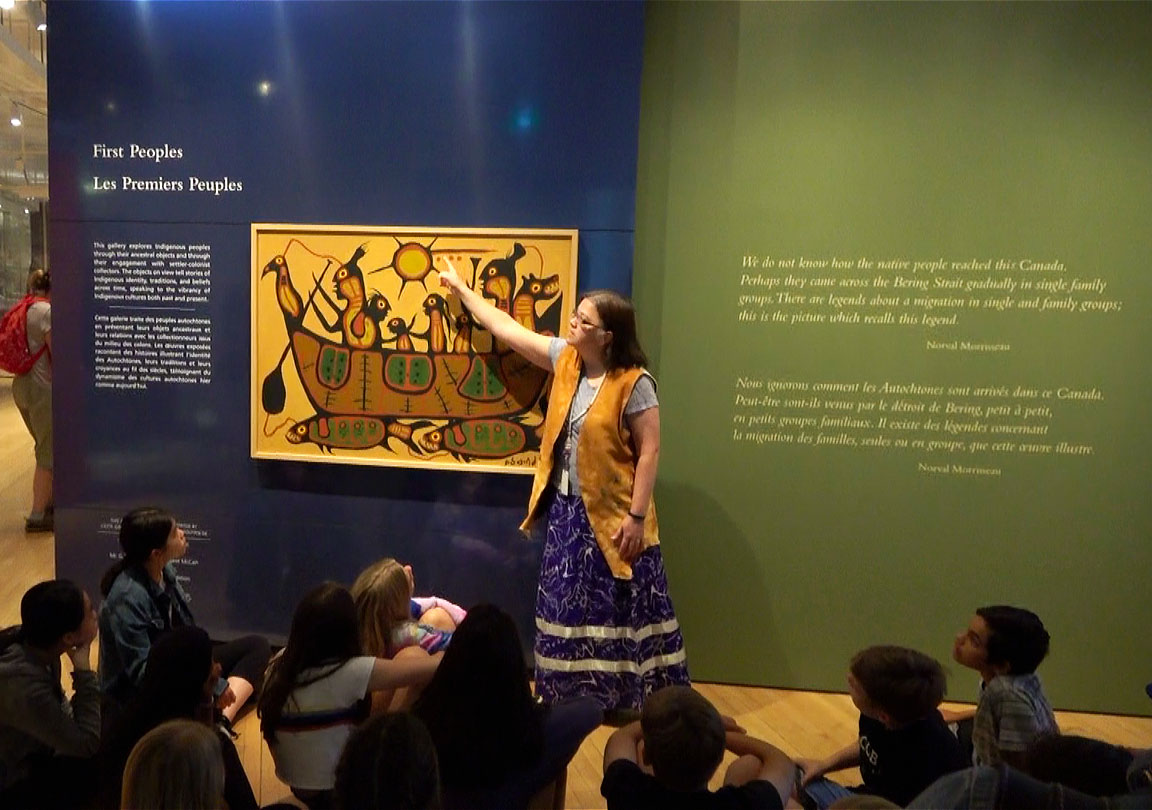An indigenous woman points at a painting. Students sit at her feet.