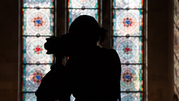 A backlit silhouette of a student with a camera, against a backdrop of stained glass windows