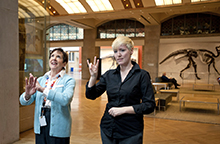 Two female sign language speakers in front of a dinosaur case
