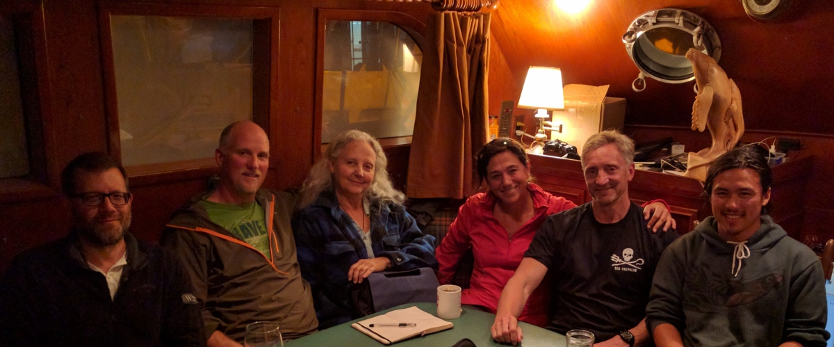 A group of 4 men & 2 women sitting around a table in a boat's cabin