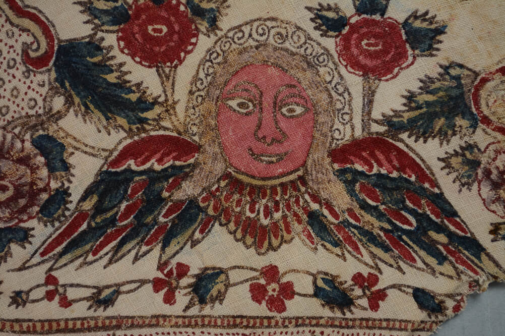 A detail of the border design of a head with face resting on a pair of outstretched angel wings.