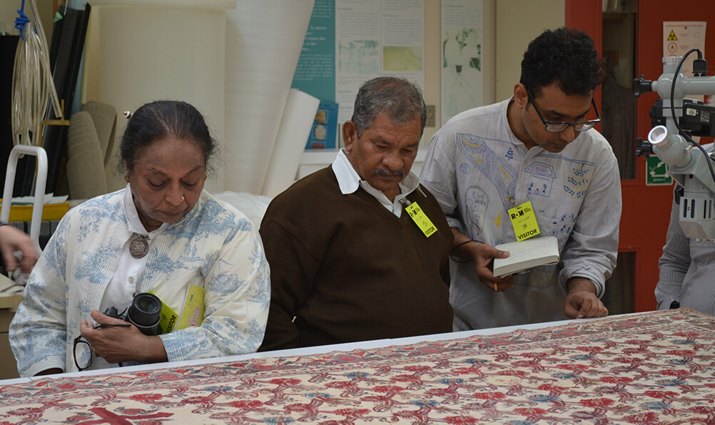 Three people, two men and a woman, examine the cope which is resting on a large table.