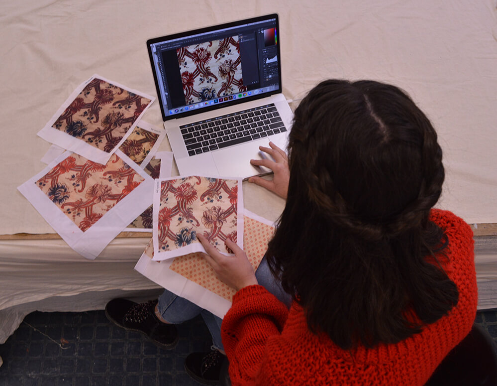 A young woman sits in front of a laptop computer surrounded by squares of patterned fabric.