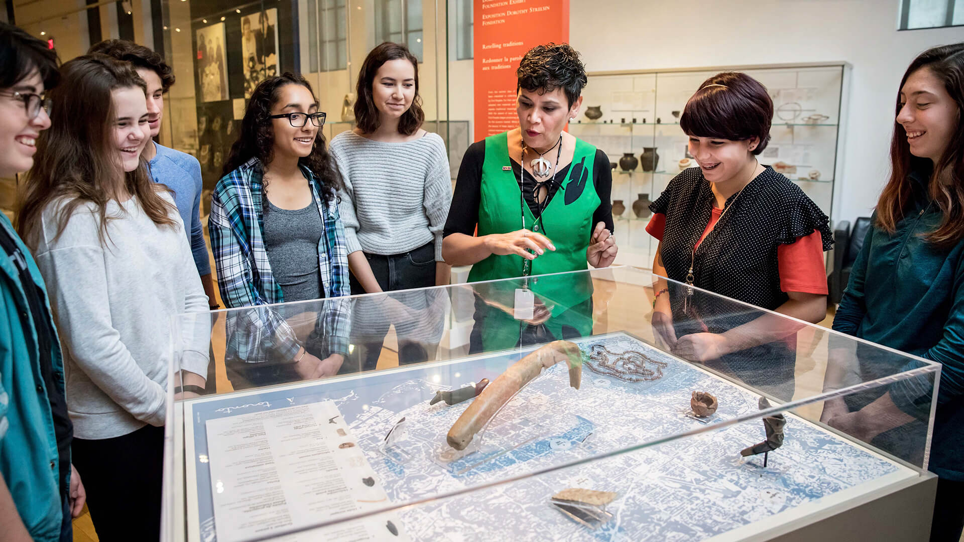 ROM staff member stands in front of a display case containing Indigenous objects with a small group of teenage students.