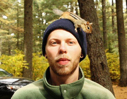 A headshot of a man standing in the woods, with a chipmunk perched on his hat.