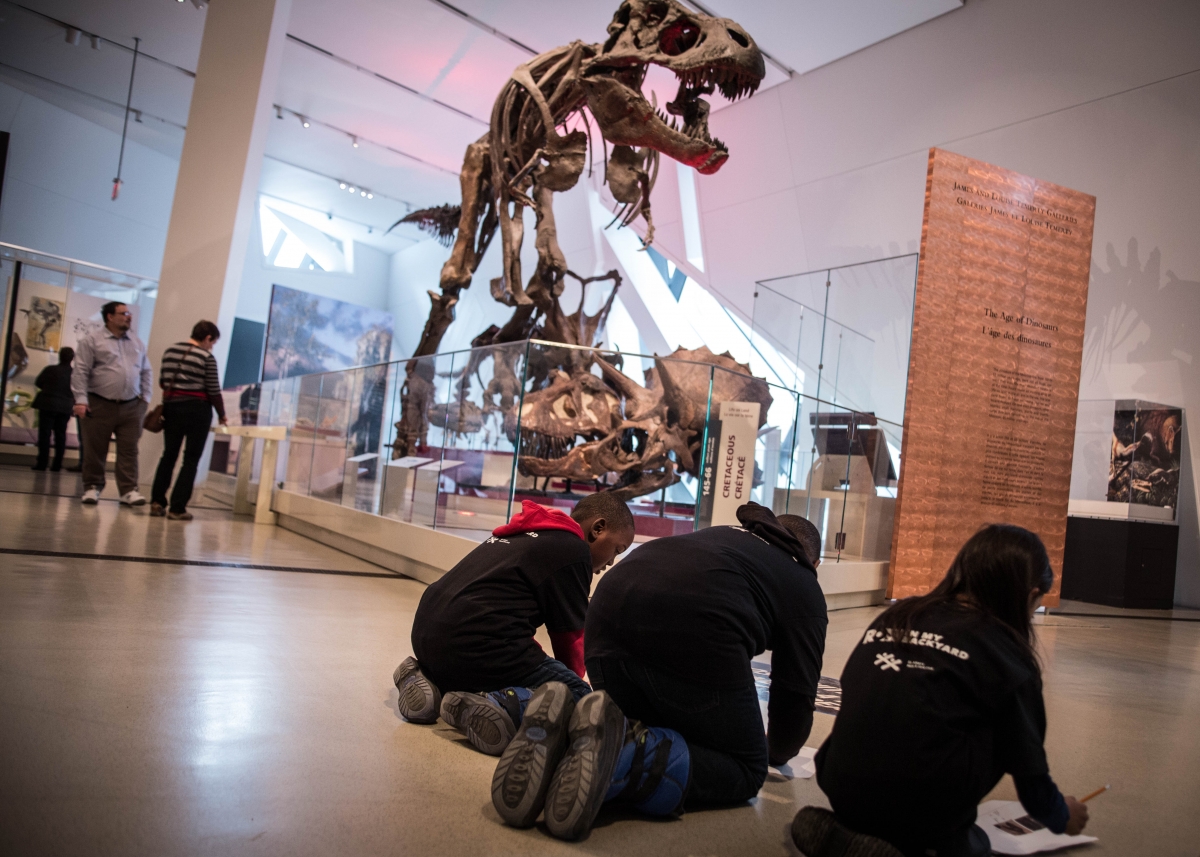 Des enfants participant au programme Le ROM dans ma cour remplissent une feuille d’activité devant un Tyrannosaurus rex au ROM