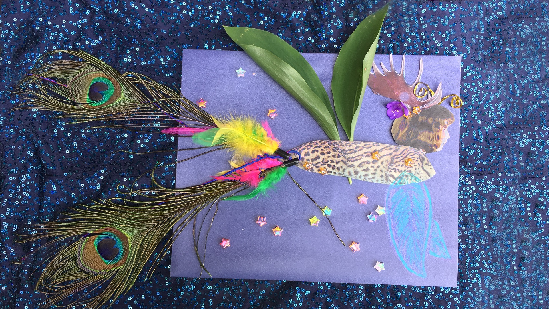 A collage, on a blue background, of a chimera with a human child’s face, moose antlers, gold antennae, blue and purple fish fins, wings made of leave, spotted ocelot body, and a tail made of colourful feathers. A flower under the antlers and star-shaped beads decorate the whole artwork.