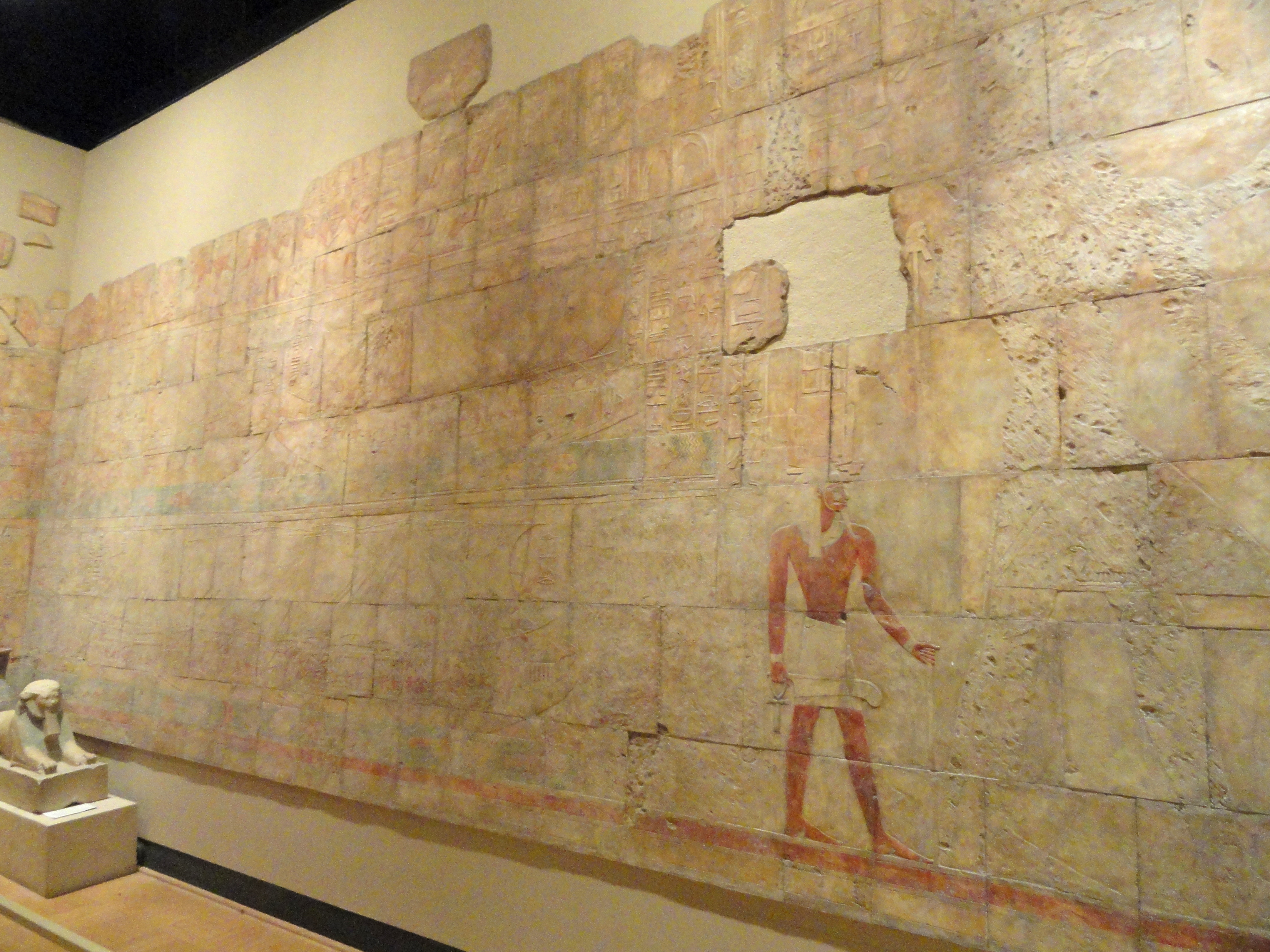 A partially-vandalized temple wall showing Queen Hatshepsut’s voyage to Punt. A small sphinx with Hatshepsut’s face sits in the lower left corner.