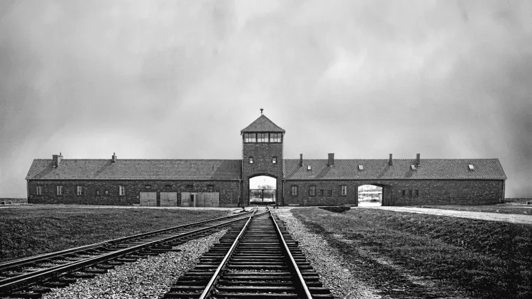 Black and white archival image showing train tracks in the foreground leading into the distance towards the entrance to the Auschwitz camp.