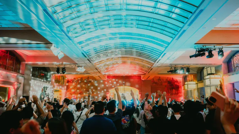 Crowd of people in a large indoor venue with a high curved glass ceiling, raising their arms toward a brightly lit stage. Colorful red and blue lighting and spotlights illuminate the space, with artwork and event lighting visible along the walls