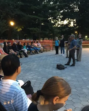 Group of people seated in a park at dusk, listening to a speaker.