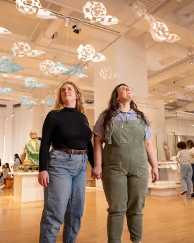 Two visitors hold hands while looking up at a hanging art installation in a museum gallery.