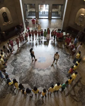 La caméra regarde de haut un groupe d'étudiants assis en cercle au fond d'une salle ronde ornée.