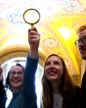 A group of people smile and hold up a magnifying glass while exploring the ornate golden ceiling of the museum rotunda.