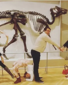 A group of people poses playfully in front of a dinosaur skeleton, arranging their bodies to spell out shapes.