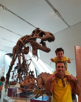 An adult carrying a smiling child on their shoulders poses beside a large Tyrannosaurus rex skeleton in a museum gallery.