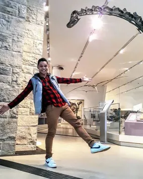 A person smiling and posing with arms outstretched in a museum gallery, standing beneath the long curved neck of a large dinosaur skeleton display.