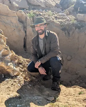 Tom Dudgeson wearing a baseball cap, sunglasses, and dark outdoor clothing crouches beside a large dinosaur skull partially embedded in rocky terrain.