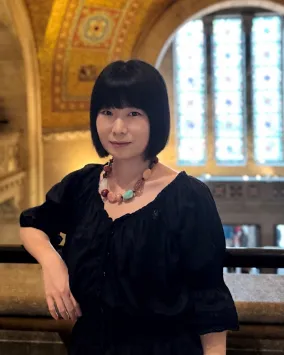 Portrait d'une femme aux cheveux noirs courts, vêtue d'un chemisier noir et portant un collier, debout à l'intérieur d'une pièce avec, en arrière-plan, une fenêtre cintrée et un plafond orné.