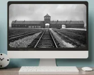 Auschwitz Virtual Exhibition A computer on a desktop shows a black-and-white image railroad tracks leading toward an arched gate beneath a peaked tower.