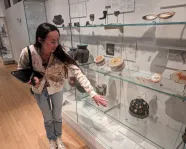 An Indigenous woman with long, dark hair and glasses stands by a museum case filled with belongings showing beadwork, quillwork, and other artistic traditions.