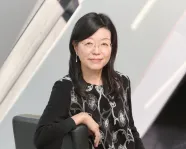 Asian woman with glasses wearing a black dress with a white floral pattern, sitting on a black chair against a modern background.
