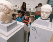 Two adults watch a smiling group of children in front of a group of marble busts