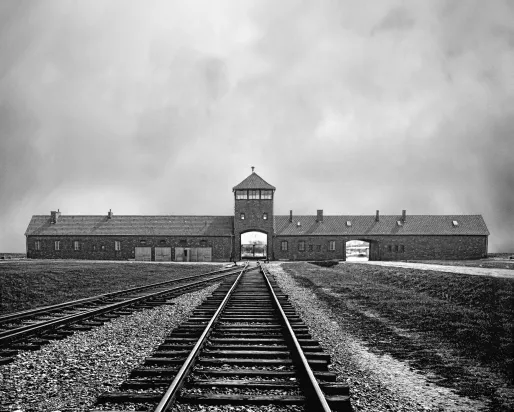 Black and white archival image showing train tracks in the foreground leading into the distance towards the entrance to the Auschwitz camp.