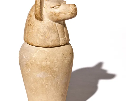 Jarre égyptienne de couleur sable dont le couvercle est sculpté en forme de tête de chacal.