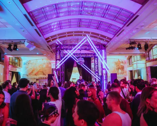 A crowd in a museum conversing. In the background is a DJ under a disco ball.