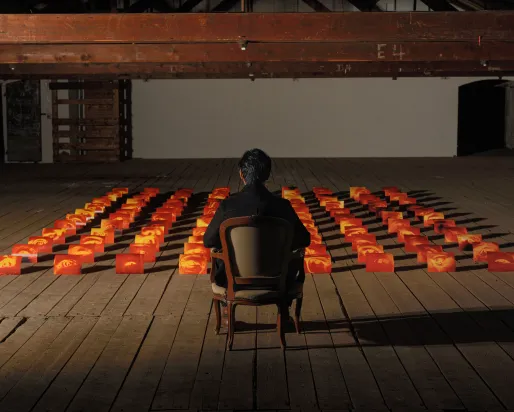 A person sits alone in a wooden chair facing an art installation of glowing orange and red blocks arranged in a grid, each block featuring a human eye, set in a dimly lit industrial-style room with exposed beams and wooden flooring.
