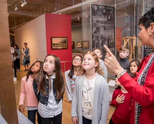 A group of children listens to a guide explaining an exhibit.