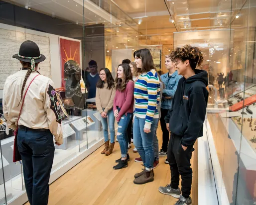 A museum educator in traditional Indigenous dress with braids and a black hat guides a group of diverse students through a history exhibit with glass display cases containing artifacts and documents.