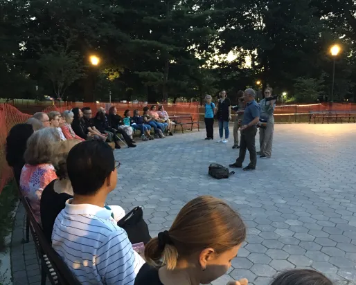 Group of people seated in a park at dusk, listening to a speaker.
