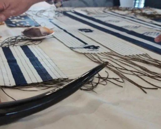 Close-up of hands working on a traditional wampum belt weaving with beads, threads, and tools laid out on a table.