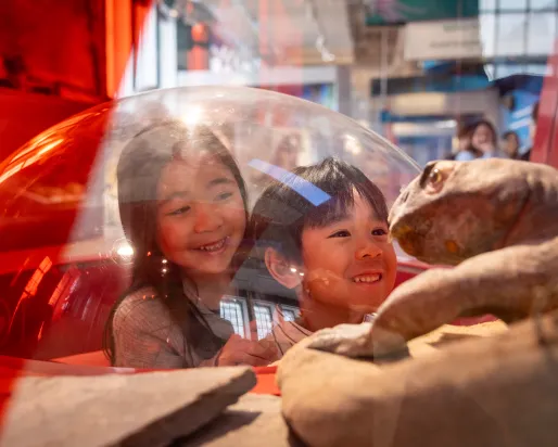 Two smiling children look through a clear dome display at a prehistoric reptile model in a museum exhibit.