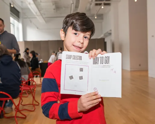 Boy in a red and blue striped sweater holding a white paper booklet with a QR code and text, indoors at an event.