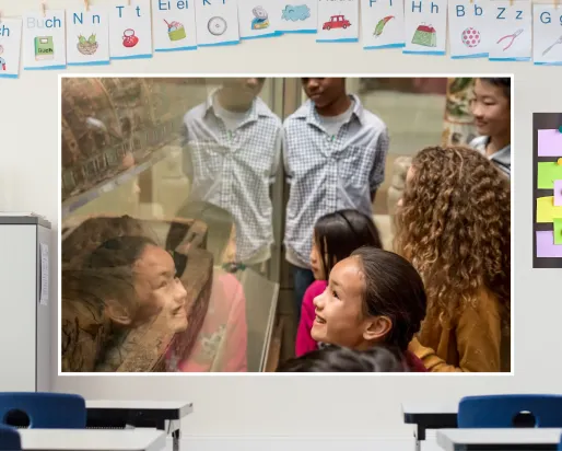 A screen in a classroom shows a scene of smiling students gathered around a museum case holding a mummified Ancient Egyptian